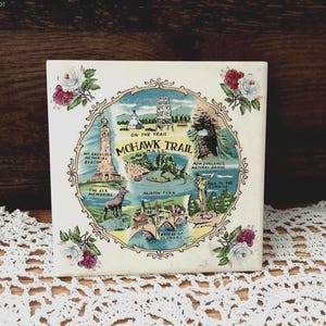 May include: A square ceramic tile featuring a colorful illustration of the Mohawk Trail in Massachusetts. The central graphic depicts landmarks like Mt. Greylock Memorial Beacon and the Bridge of Flowers, framed by floral accents in the corners.