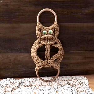 May include: A tan macrame wall hanging featuring a stylized creature design with two round metal rings at the top and bottom. The creature has two green plastic eyes and a knotted cord body structure that connects the upper and lower metal rings.