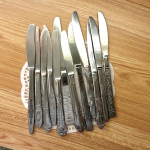 May include: A collection of eleven silver-toned metal dinner knives arranged in a fan shape. Each knife features a distinct handle design, ranging from smooth and plain to intricate floral or geometric embossed patterns on the metal surfaces.