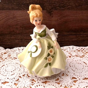 May include: A porcelain figurine of a woman in a yellow dress, holding a white hat with a green ribbon. The dress is adorned with floral accents. The figurine has blonde hair and is set on a decorative base.