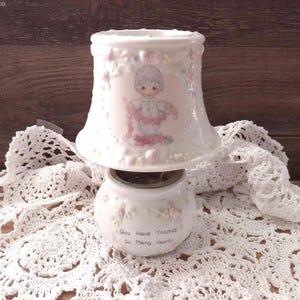 May include: A white ceramic candle warmer featuring a bell-shaped shade with a pastel illustration of a child holding a garland of pink flowers. The base is a rounded pot with the text "You Have Touched So Many Hearts" printed in black lettering.