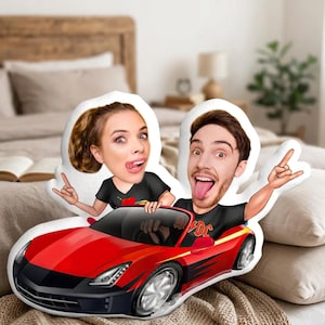 Puede incluir: Un cojín de diseño personalizado con una pareja de dibujos animados en un coche deportivo rojo. La mujer saca la lengua y hace un gesto de rock. El hombre también saca la lengua y hace un gesto de rock.