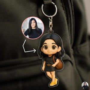 以下が含まれることがあります： 黒髪で黒いドレスと黄色の靴を履き、茶色のバッグを持った人物の漫画のキーホルダー。人物の写真が、その人物の上のピンク色の円の中にあります。キーホルダーはシルバーのキーリングに付いています。