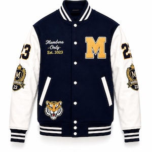 Könnte beinhalten: Eine marineblaue Collegejacke mit weißen Ärmeln, einem großen goldenen Buchstaben M auf der Brust, einem Tigerkopf-Aufnäher und dem Text Members Only Est. 2023. Die Ärmel zeigen die Zahl 23 sowie ein Wappen mit einer Krone und dem Buchstaben M.