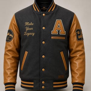 Puede incluir: Una chaqueta universitaria gris carbón con mangas de cuero color canela, con una letra A grande en el pecho, el texto Make Your Legacy, un gráfico de montaña en la manga derecha y el número 23 en la manga izquierda con ribetes dorados y negros.