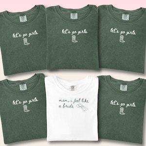 May include: Six folded t-shirts. Five are olive green, featuring the text "let's go girls" and a cowboy boot illustration. One white shirt has the text "man, I feel like a bride" with a hat graphic.