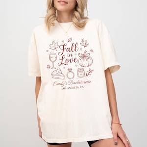 May include: An off-white short-sleeve t-shirt featuring a dark red graphic print with the text Fall in Love, Emily's Bachelorette, and LOS ANGELES, CA. The design includes illustrations of a wine glass, pumpkin, pie slice, ring, and candle.
