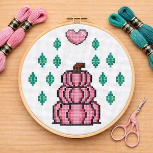 Könnte beinhalten: Ein Kreuzstich-Stickrahmen mit einem rosa Kürbismotiv, einem Herz und grünen Akzenten. Rosa und türkisfarbene Stickgarne und eine rosa Schere umgeben den Rahmen und bilden eine handwerkliche Komposition.