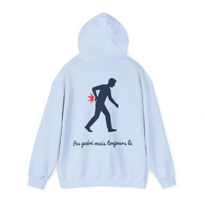 May include: Light blue hoodie with a hood and long sleeves. The back features a black silhouette of a person walking with a red pain symbol, and the text "Pas guéri mais toujours là".