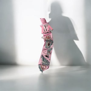 May include: A long, pink satin necktie adorned with various embellishments including a small pink jacket cutout, a safety pin with pearls, frayed denim fabric patches, and a heart-shaped charm. The tie features a smooth, glossy finish.