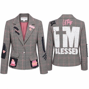 May include: A gray plaid blazer with pink accents and patches. The front features a pink flower pin and patches with text and graphics. The back of the blazer has the text "I'M BLESSED" in white and pink lettering. The blazer has a pink lining.