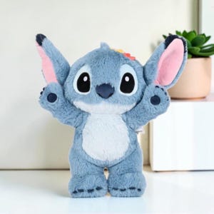 Puede incluir: Un peluche azul con grandes orejas puntiagudas con interior rosa, nariz azul oscuro y grandes ojos negros. El personaje tiene una mancha azul claro en el vientre, brazos levantados y pies peque&ntilde;os con almohadillas azul oscuro en tela suave.