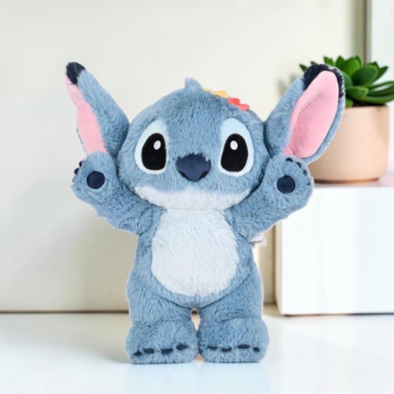 Puede incluir: Un peluche azul con grandes orejas puntiagudas con interior rosa, nariz azul oscuro y grandes ojos negros. El personaje tiene una mancha azul claro en el vientre, brazos levantados y pies peque&ntilde;os con almohadillas azul oscuro en tela suave.