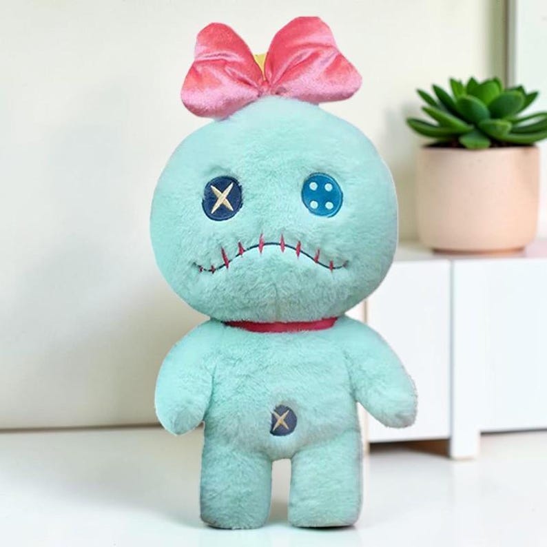 Puede incluir: Una mu&ntilde;eca de peluche con suave pelaje verde menta, que presenta un gran lazo rosa en la parte superior de la cabeza. La cara tiene un ojo de bot&oacute;n azul oscuro con una costura en X, un ojo de bot&oacute;n azul claro con cuatro agujeros y una boca roja cosida. Un cuello rojo y un bot&oacute;n azul en el torso completan el dise&ntilde;o.