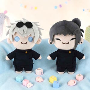 Könnte beinhalten: Zwei Plüschpuppen von Anime-Charakteren, eine mit weißen Haaren und schwarzer Sonnenbrille, die andere mit schwarzen, hochgebundenen Haaren. Beide Puppen tragen schwarze Uniformen. Kleine, bunte Blöcke und Figuren sind auf einer blauen Oberfläche um sie herum verstreut.