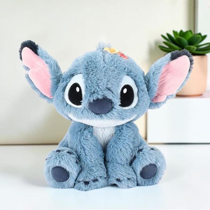 Puede incluir: Un peluche del personaje Stitch, con pelaje azul suave, orejas grandes y puntiagudas con interior rosa, ojos negros de gran tama&ntilde;o, nariz azul oscuro y un peque&ntilde;o detalle de flor amarilla y roja en la parte superior de la cabeza, en posici&oacute;n sentada.