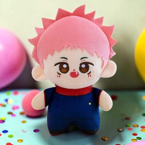 Puede incluir: Una muñeca de peluche con un peinado rosa puntiagudo, ojos marrones y una pequeña boca roja. La muñeca lleva un atuendo azul marino con detalles en rojo. La muñeca es un juguete suave y lindo.