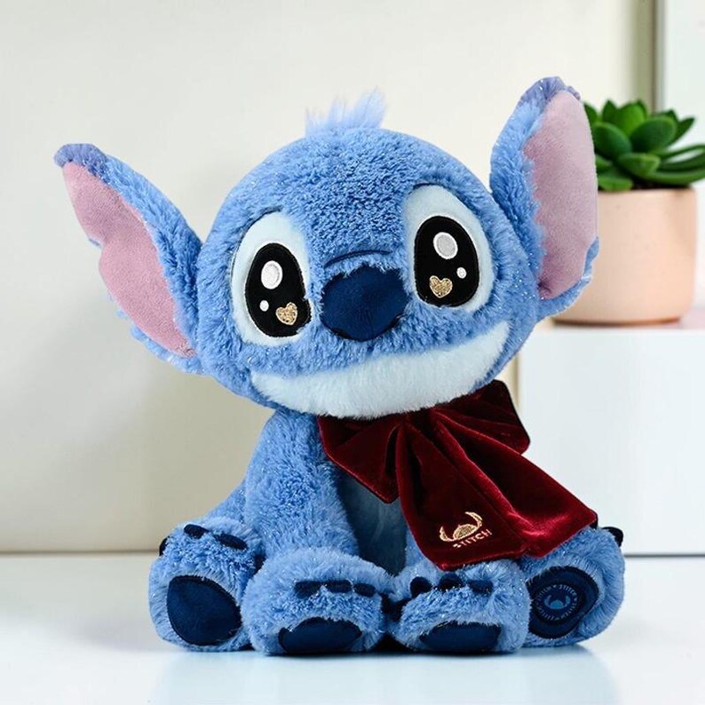 Puede incluir: Un peluche azul con orejas grandes, interiores rosados y ojos negros sobredimensionados con detalles de corazones dorados. El personaje lleva una bufanda de terciopelo rojo oscuro con bordado dorado que dice STITCH y almohadillas azul oscuro.