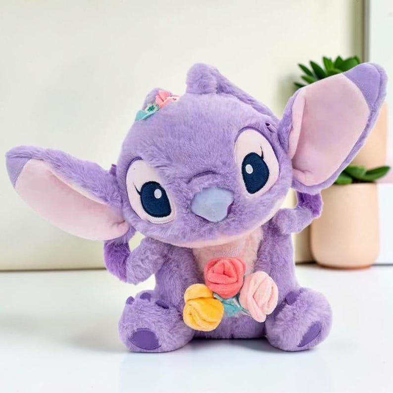 Puede incluir: Un juguete de peluche con pelaje color lavanda, grandes orejas puntiagudas con secciones internas rosa claro y ojos azul oscuro. El personaje presenta un peque&ntilde;o grupo de rosas de tela suave en tonos rosa, amarillo y melocot&oacute;n en el pecho.