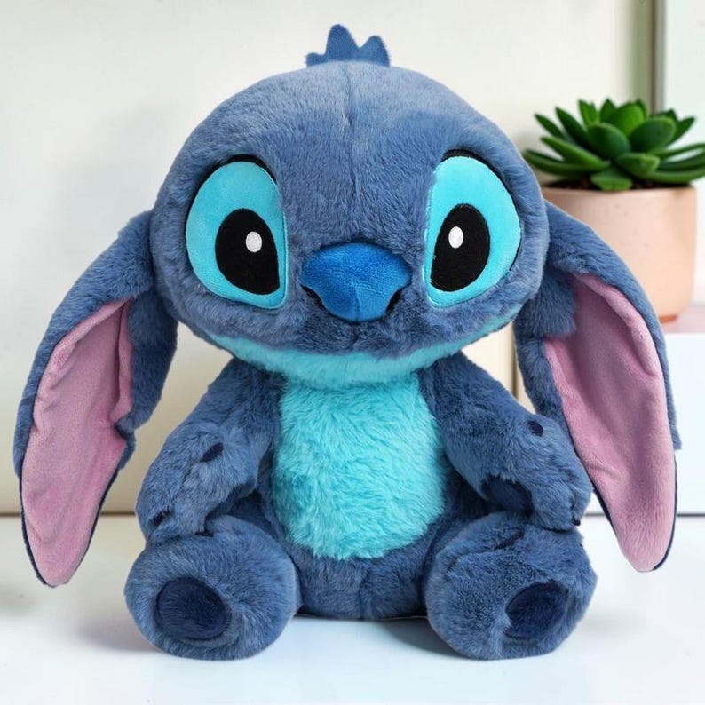 Puede incluir: Un peluche con pelaje azul suave, grandes ojos ovalados con pupilas negras e iris azul claro, una nariz azul oscuro y orejas largas con forro interior rosa. El personaje tiene una mancha azul claro en el pecho y almohadillas azul oscuro.