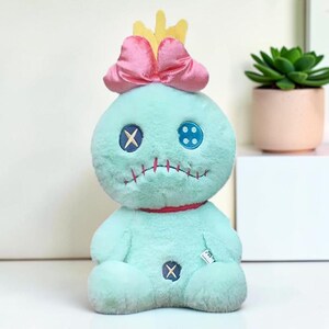 Puede incluir: Una mu&ntilde;eca de peluche con suave pelaje verde menta, con un gran lazo rosa y una corona amarilla en la cabeza. La cara tiene un ojo de bot&oacute;n azul oscuro con una costura en X y un ojo de bot&oacute;n azul claro, adem&aacute;s de una boca roja cosida.