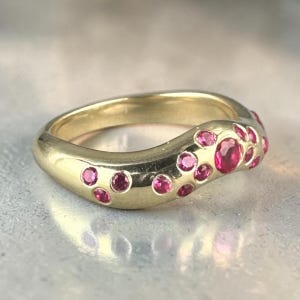 May include: A gold ring with a curved design, featuring numerous round, bright pink gemstones. The ring's band is smooth and polished, reflecting light. The gemstones are set in a cluster pattern along the top of the ring.