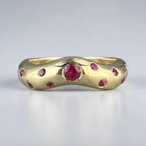 May include: A gold-colored ring with a wavy design, set with multiple small, round, red gemstones. The ring features a larger, central red gemstone. The ring's smooth, polished surface reflects light, highlighting the gemstones.