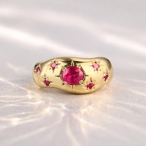May include: A gold ring featuring a large, round, red gemstone at its centre, encircled by smaller, star-shaped red gemstones. The ring has a smooth, rounded design.