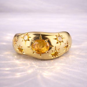 May include: A gold ring featuring a central oval yellow gemstone, surrounded by star-shaped embellishments and smaller yellow gems. The ring's surface is smooth and polished, with a curved design. The ring is a statement piece.