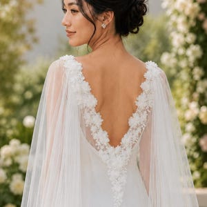May include: A white floor-length gown featuring a deep V-neckline on the back, adorned with intricate white floral appliqués along the edges. The dress includes a sheer, flowing tulle cape that drapes from the shoulders, creating a soft silhouette.