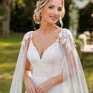 Puede incluir: Un vestido de novia blanco largo hasta el suelo con escote en V, detalles de encaje intrincado en el corpiño y una capa de tul transparente sujeta a los hombros con aplicaciones florales de encaje. El vestido tiene una silueta ajustada.