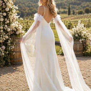 Peut inclure: Une robe de mariée blanche longue jusqu'au sol avec un dos en V profond, de fines bretelles et des manches amovibles en tulle transparent ornées de volants aux épaules, tombant en longs pans fluides de tissu léger.