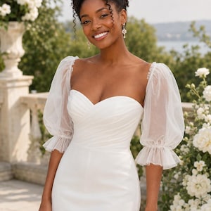 Puede incluir: Un vestido blanco largo hasta el suelo con escote corazón, cuerpo plisado y mangas abullonadas transparentes de tul. Las mangas están fruncidas a la altura de los codos con puños de volantes, lo que crea una silueta muy definida.