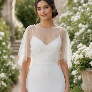 May include: A white sleeveless dress featuring a V-neckline and a sheer tulle capelet overlay. The capelet is adorned with scattered small white pearls and delicate floral appliqués, creating a soft, layered effect over the bodice.