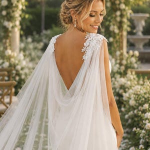 Puede incluir: Un vestido de novia blanco largo hasta el suelo con espalda descubierta, cuerpo sin mangas y una capa de tul transparente sujeta a los hombros con intrincados apliques de encaje floral blanco que caen elegantemente por la espalda.