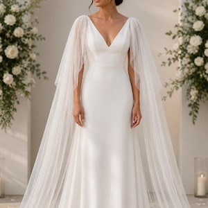 Peut inclure: Robe de mariée blanche avec un décolleté en V et une jupe longue. La robe est ornée de manches en forme de cape transparente qui se prolongent en une longue traîne. La mariée se tient devant des compositions florales.
