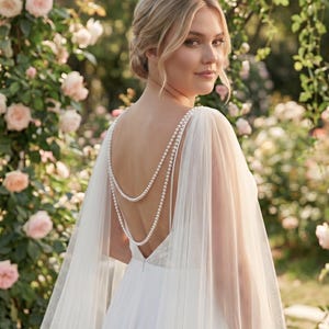 Puede incluir: Vestido de novia blanco con capa transparente y detalles de perlas en la espalda. La novia está en un jardín con rosas rosas y vegetación. El vestido tiene la espalda baja y una falda fluida.
