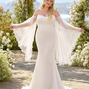 Puede incluir: Un vestido blanco largo hasta el suelo, sin tirantes, con escote corazón y cuerpo ajustado que se ensancha en una silueta de trompeta. El vestido incluye mangas de tul transparente desmontables con puños anchos y fluidos con volantes.