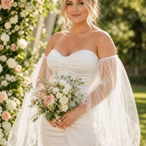 K&ouml;nnte beinhalten: Ein wei&szlig;es tr&auml;gerloses Kleid mit herzf&ouml;rmigem Ausschnitt und transparenten, flie&szlig;enden T&uuml;ll&auml;rmeln, die &uuml;ber die Schultern fallen und ger&uuml;schte Kanten haben. Das Kleid wird mit einem Strau&szlig; aus wei&szlig;en und blassrosa Rosen kombiniert.