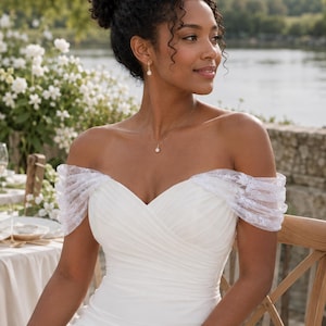 Puede incluir: Un vestido de novia blanco con hombros descubiertos, escote corazón, cuerpo drapeado y mangas cortas de encaje floral transparente. El diseño cuenta con un corpiño ajustado que se extiende hacia una falda de silueta fluida.