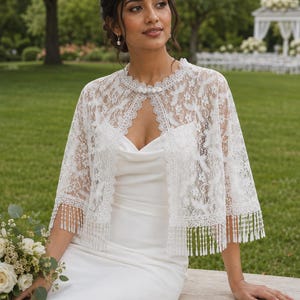 Puede incluir: Una chaqueta bolero de encaje blanco con un intrincado patrón floral, un escote festoneado y una fila de tres botones tipo perla en el cuello. El dobladillo está acabado con largos y delicados flecos de cuentas que cuelgan de los bordes.
