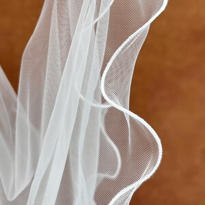 K&ouml;nnte beinhalten: Ein transparenter wei&szlig;er Schleier aus feinem Netzstoff mit einem markanten, wellenf&ouml;rmigen, gebogten Rand, der mit einer d&uuml;nnen, soliden wei&szlig;en Kante versehen ist. Das leichte Material f&auml;llt in weichen, flie&szlig;enden Falten.