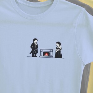 Puede incluir: Camiseta azul claro con una ilustración minimalista en blanco y negro de una pareja frente a una chimenea. El diseño muestra a un hombre y una mujer con ropa de época. La camiseta está colgada en una percha con otras.