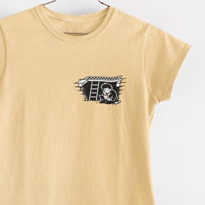 May include: A light yellow t-shirt with a small black and white graphic on the chest. The graphic depicts a person looking out from a structure with a ladder. The shirt is hanging from a metal hanger against a white background.
