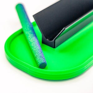 May include: A bright green tray with a rounded rectangular shape. A textured, cylindrical object with blue and green hues rests on the tray, along with a black, angled object. The objects are set against a white background.