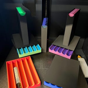 May include: A collection of 3D-printed organizers in various colors. The organizers include black stands with colored trays in green, blue, purple, and pink. A red tray holds a glass tube. The items are on a dark surface.