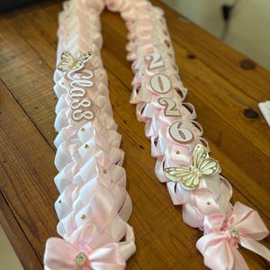 May include: Two pink graduation leis with white accents, bows, and butterfly embellishments. One lei reads "Class" and the other displays "2026". Each lei is adorned with small gold-colored studs and a large bow with a rhinestone center.