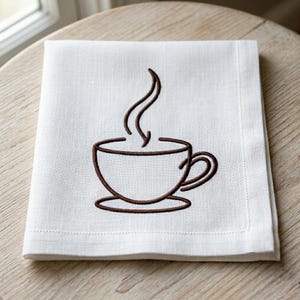 Könnte beinhalten: Ein weißes Leinen-Servietten mit einer braunen Stickerei einer Kaffeetasse mit aufsteigendem Dampf. Die Serviette ist gefaltet und liegt auf einer Holzoberfläche. Das Design ist schlicht und elegant.