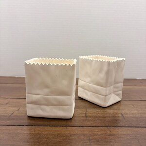 May include: Two off-white ceramic containers shaped like small paper bags. Each vessel features a square base, a crinkled texture on the sides, and a serrated, zigzag-cut top edge. The containers have a matte finish and a structured, upright form.