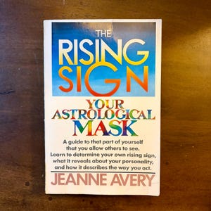 May include: A paperback book titled The Rising Sign: Your Astrological Mask by Jeanne Avery. The cover features a blue gradient background with large orange text, followed by multicolored text and a white section with black descriptive text.